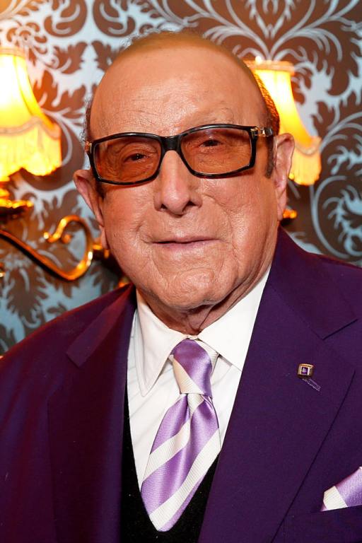 Clive Davis wearing a purple suit and purple and white striped tie wearing amber colored glasses.