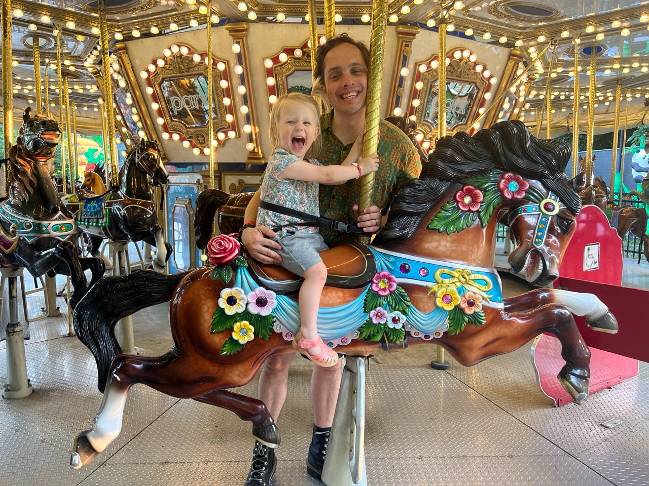 Photo of Kristen's husband Paul and daughter Trudy riding the carousel at Franklin Square. Both smiling with Trudy happily sitting on the horse and Paul standing behind her.